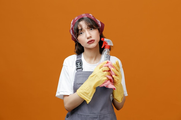 Traurige junge Putzfrau mit einheitlichen Gummihandschuhen und Bandana mit Reinigungsmittel und Stoffstaubtuch, die isoliert auf orangefarbenem Hintergrund in die Kamera blickt