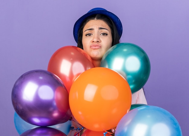 Traurige junge schöne Frau mit Partyhut, die hinter Luftballons steht, isoliert auf blauer Wand