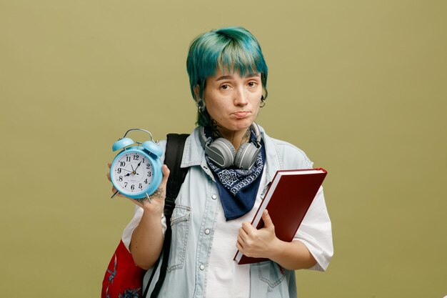 Traurige junge Studentin mit Kopfhörern und Bandana am Hals und Rucksack mit Notizbuch, das in die Kamera schaut und den Wecker isoliert auf olivgrünem Hintergrund zeigt