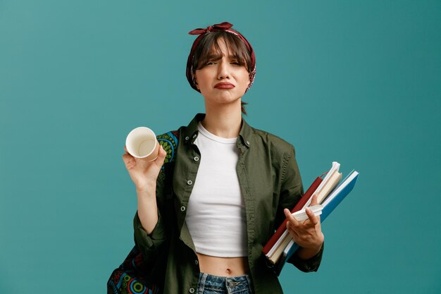 Traurige junge Studentin mit Kopftuch und Rucksack mit Notizblöcken aus Papierkaffeetasse und ihrer Kappe, die in die Kamera blickt und eine leere Kaffeetasse isoliert auf blauem Hintergrund zeigt