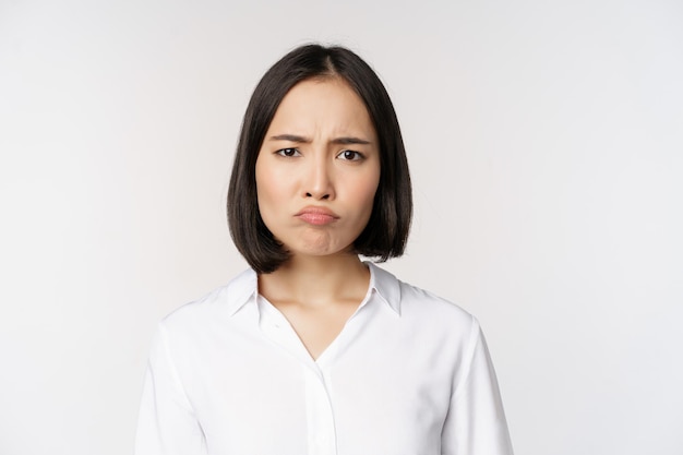 Traurige und düstere junge asiatische Frau, die das Gesicht verzieht und die Stirn runzelt, macht schmollendes Gesicht auf weißem Hintergrund