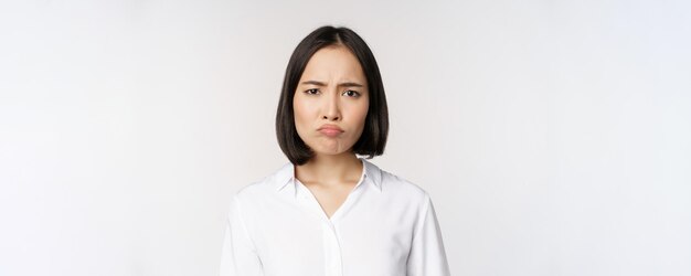 Traurige und düstere junge asiatische Frau, die das Gesicht verzieht und die Stirn runzelt, macht schmollendes Gesicht auf weißem Hintergrund