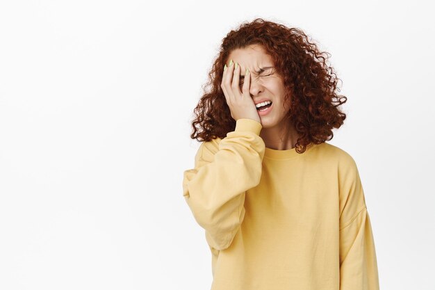 Traurige und enttäuschte rothaarige Frau facepalm, verärgert aussehend, beunruhigt durch schlechte beunruhigende Nachrichten, frustriert vor weißem Hintergrund stehend