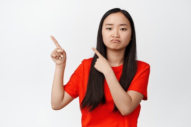 Traurige und launische Asiatin, die mit den Fingern auf die obere linke Ecke zeigt, schmollend und stirnrunzelnd verärgert, in rotem T-Shirt auf Weiß stehend