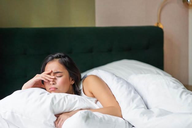 Traurige und verzweifelte junge Koreanerin, die mit Kissen und warmer Bettdecke in ihrem Bett liegt, fühlt sich unwohl und runzelt die Stirn