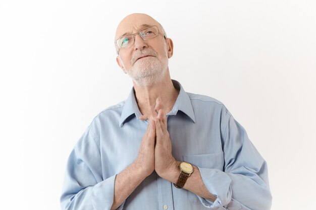 Trauriger alter reifer Mann in Brille und blauem Hemd mit hoffnungsvollem Gesichtsausdruck, Hände im Gebet zusammengedrückt, auf das Beste hoffend, während er sich Schwierigkeiten, Stress oder Problemen gegenübersieht