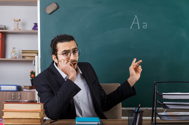 Trauriger gepackter männlicher Lehrer mit Brille zeigt an der Tafel, die am Tisch mit Schulwerkzeugen im Klassenzimmer sitzt
