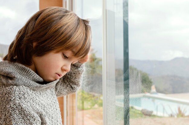 Trauriger Junge der Seitenansicht, der durch Fenster steht