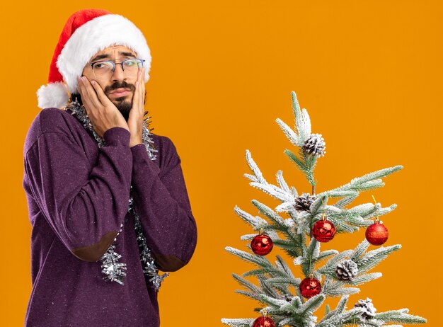 Trauriger junger hübscher Kerl, der nahe Weihnachtsbaum steht, der Weihnachtshut mit Girlande am Hals trägt Hände auf Wangen lokalisiert auf orange Hintergrund setzt