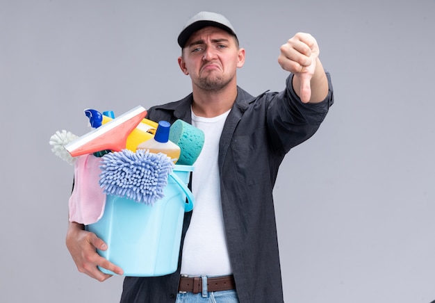 Trauriger junger hübscher reinigungsmann, der t-shirt und kappe hält, die eimer mit reinigungswerkzeugen hält, der daumen unten lokalisiert auf weißer wand zeigt