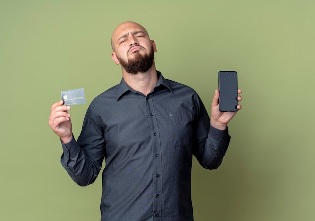 Trauriger junger kahlköpfiger Callcenter-Mann, der Handy und Kreditkarte mit geschlossenen Augen lokalisiert auf olivgrüner Wand hält