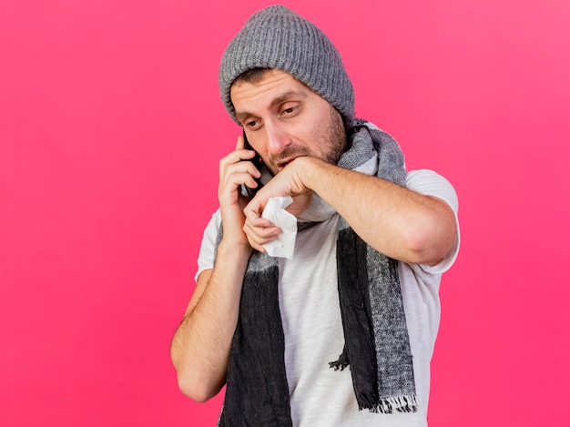 Trauriger junger kranker Mann, der Wintermütze mit Schal trägt, spricht am Telefon, das Serviette hält und Kinn mit Hand lokalisiert auf rosa Hintergrund abwischt