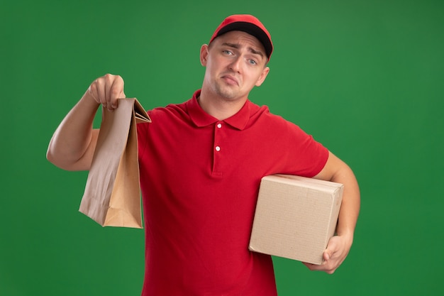 Trauriger junger Lieferbote, der Uniform und Kappe hält, die Papiernahrungsmittelpaket mit Kasten lokalisiert auf grüner Wand hält