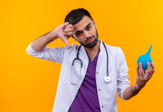 Trauriger junger männlicher Arzt, der das medizinische Kleid des Stethoskops trägt, das Einlauf seinen Daumen unten auf lokalisiertem gelbem Hintergrund hält