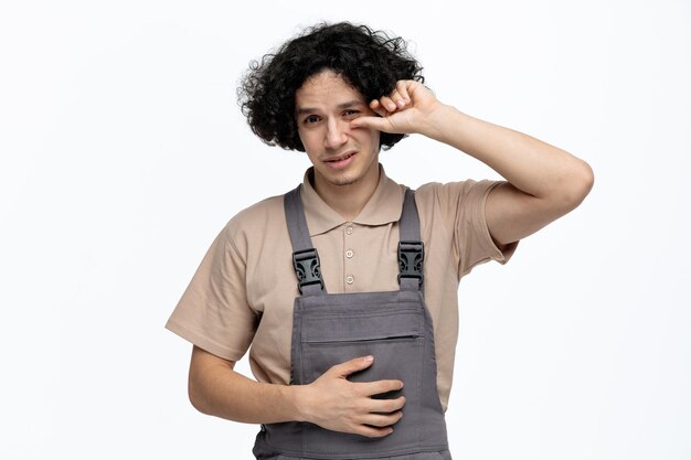 Trauriger junger männlicher Bauarbeiter in Uniform, der in die Kamera blickt und die Hand auf den Bauch hält, die auf sein Auge zeigt, isoliert auf weißem Hintergrund