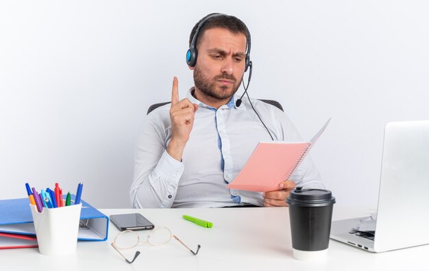 Trauriger junger männlicher Callcenter-Betreiber mit Headset am Tisch sitzend mit Bürowerkzeugen, die ein Notebook halten und lesen