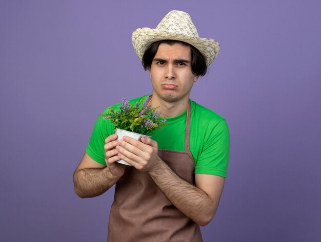 Trauriger junger männlicher Gärtner in der Uniform, die Gartenhut hält, der Blume im Blumentopf hält, der auf Purpur lokalisiert wird