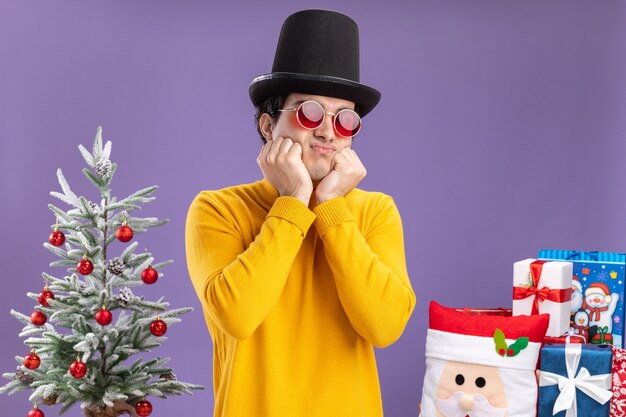 Trauriger junger Mann im gelben Rollkragenpullover und in der Brille, die schwarzen Hut tragen, der neben einem Weihnachtsbaum steht und über lila Hintergrund präsentiert