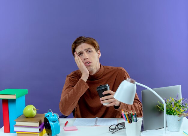 Trauriger junger studend Junge, der am Schreibtisch mit Schulwerkzeugen sitzt, die Tasse Kaffee halten, die Hand auf Wange auf lila hält