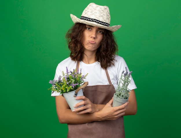 Trauriger junger weiblicher Gärtner in der Uniform, die Gartenhut trägt und Blumen in Blumentöpfen hält und kreuzt