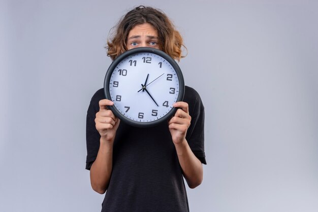 Trauriger Kerl mit langen Haaren im schwarzen T-Shirt, das eine Wanduhr auf weißer Wand hält