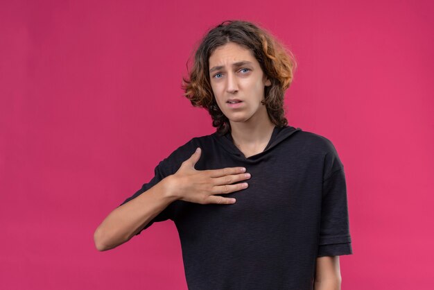 Trauriger Kerl mit langen Haaren im schwarzen T-Shirt legte seine Hand auf Brust auf rosa Wand