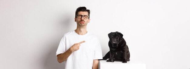 Kostenloses Foto trauriger und düsterer haustierbesitzer, der auf seinen schwarzen pug-hund zeigt und schluchzend gegen weißen bac steht