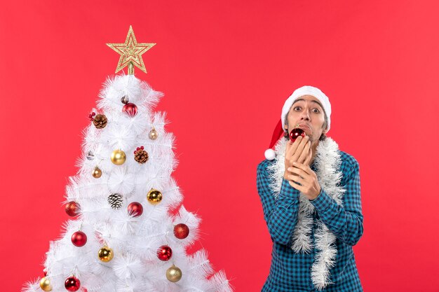 Trauriger unglücklicher emotionaler junger Mann mit Weihnachtsmannhut in einem blauen gestreiften Hemd und in der Dekoration haltend