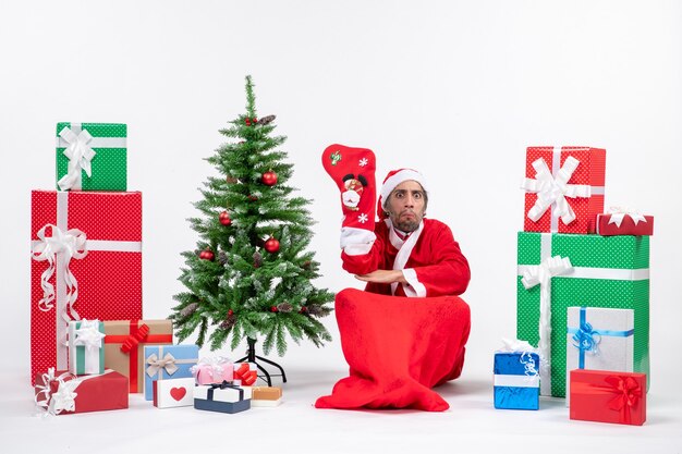 Trauriger Weihnachtsmann, der auf dem Boden sitzt und Weihnachtssocke in der Nähe von Geschenken und geschmücktem Neujahrsbaum auf weißem Hintergrund trägt