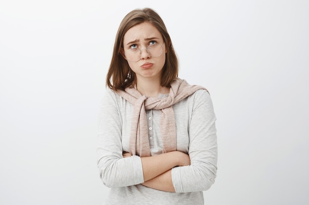 Trauriges, düsteres, beleidigtes, dummes Mädchen in Brille und süßem Outfit, das die Lippen faltet, die Stirn runzelt und aufschaut, während es sich darüber beschwert, beleidigt über das grausame und unfaire Leben über der grauen Wand zu jammern