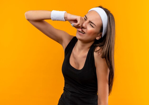 Trauriges junges hübsches sportliches Mädchen, das Stirnband und Armband weinend trägt, die Hand auf Gesicht mit geschlossenen Augen lokalisiert auf orange Raum setzen