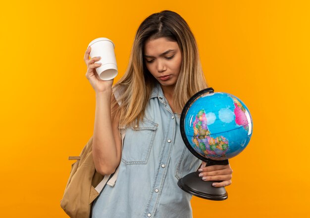 Trauriges junges hübsches Studentenmädchen, das Rückentasche hält, die Plastikkaffeetasse und Globus hält, lokalisiert auf orange Wand