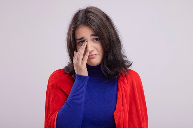 Trauriges junges kaukasisches Superheldenmädchen im roten Umhang, das Hand auf die Nase legt, die auf weißer Wand lokalisiert wird