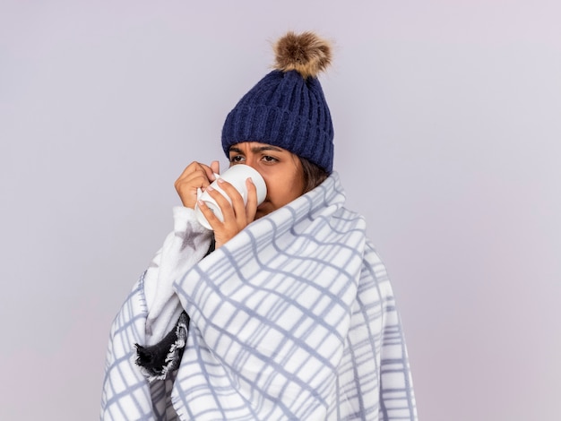 Trauriges junges krankes Mädchen, das Seite betrachtet, die Wintermütze mit Schal wickelt, der in den karierten trinkenden Tee eingewickelt wird, lokalisiert auf Weiß