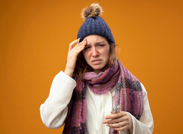Trauriges junges krankes Mädchen, das weiße Robe und Wintermütze mit Schal hält Spritze mit Pillen trägt Hand auf Stirn lokalisiert auf orange Wand