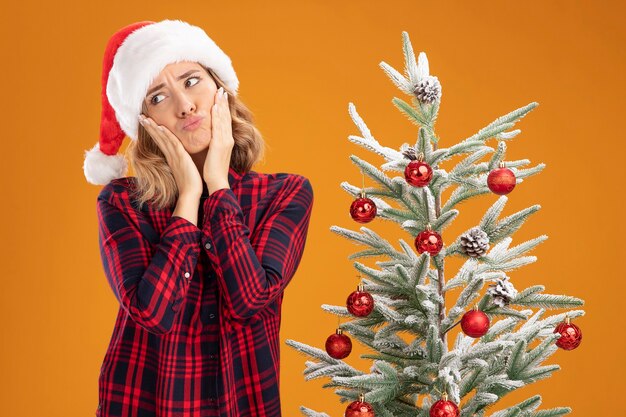 Kostenloses Foto trauriges junges schönes mädchen, das in der nähe des weihnachtsbaums mit weihnachtsmütze steht und die hände auf die wangen legt, isoliert auf orangefarbenem hintergrund