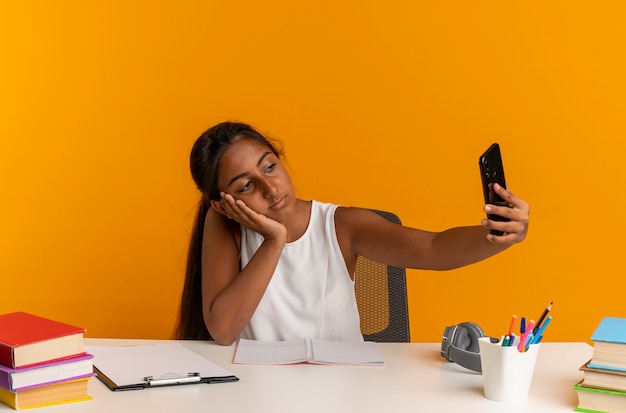 Trauriges junges Schulmädchen, das am Schreibtisch mit Schulwerkzeugen sitzt, die Kopf auf Hand setzen und Selfie lokalisiert auf orange Wand nehmen