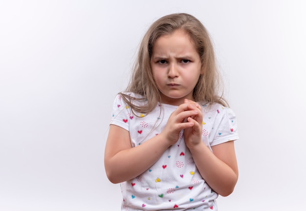 Trauriges kleines Schulmädchen, das weißes T-Shirt hält Hände zusammen auf lokalisiertem weißem Hintergrund hält