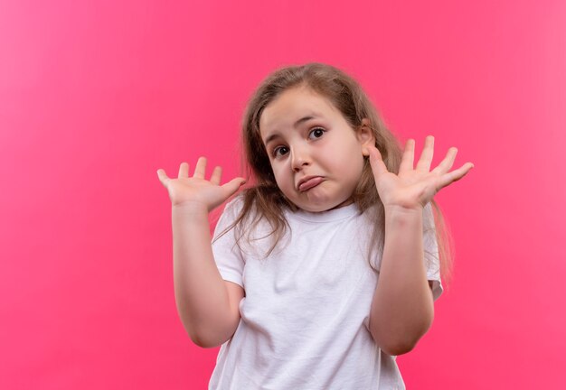 Trauriges kleines Schulmädchen, das weißes T-Shirt trägt, hob Hände auf lokalisiertem rosa Hintergrund