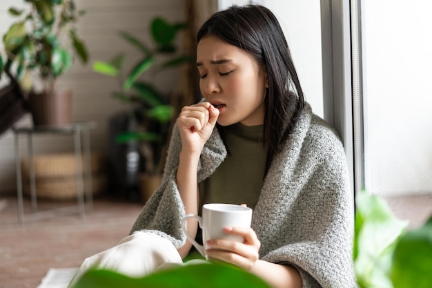 Trauriges krankes asiatisches Mädchen, das während einer Covid-Grippe in Selbstquarantäne bleibt und zu Hause mit einer Tasse sitzt ...