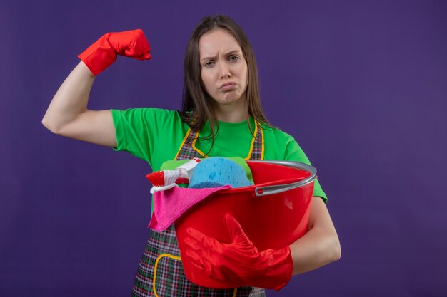 Trauriges putzendes junges Mädchen, das Uniform in Handschuhen hält, die Reinigungswerkzeuge halten, die starke Geste auf lila Hintergrund tun