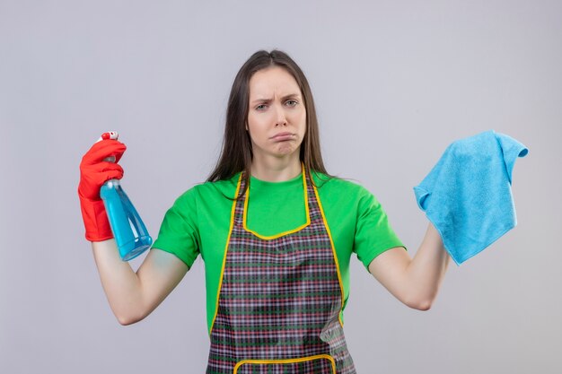 Trauriges putzendes junges Mädchen, das Uniform in roten Handschuhen hält, die Reinigungsspray und Lappen auf lokalisiertem weißem Hintergrund halten