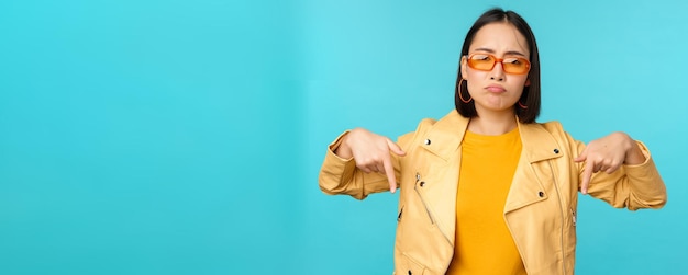 Trauriges und düsteres chinesisches Mädchen mit Sonnenbrille zeigt mit den Fingern nach unten und Grimassen sieht elend aus und enttäuschte Gefühle des Bedauerns, die über blauem Hintergrund stehen