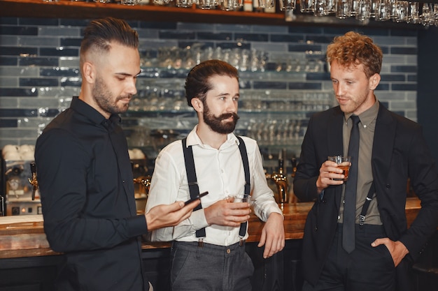 Treffen mit den besten Freunden. Drei glückliche junge Männer in Freizeitkleidung sprechen und trinken Bier, während sie zusammen in der Bar sitzen. Mann hält ein Telefon in seinen Händen.