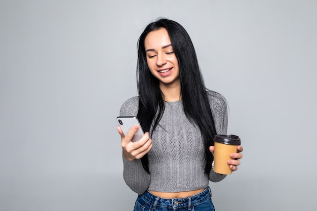 Trendige junge Frau, die auf einem Handy plaudert, während sie eine Tasse Kaffee zum Mitnehmen in ihrer anderen Hand hält, lokalisiert auf grauer Wand