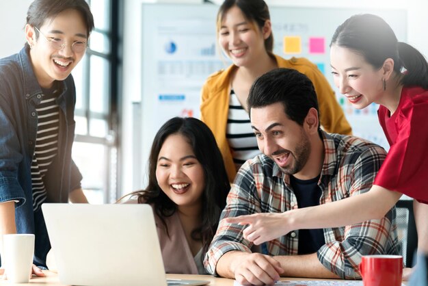 Trendiger asiatischer kreativer Hipster-Freund, der lächelt, während er bei einem ungezwungenen Treffen mit einer Gruppe von Freunden sitzt, arbeitet an einem neuen Erfolgsprojekt, während er digitale Laptops verwendet