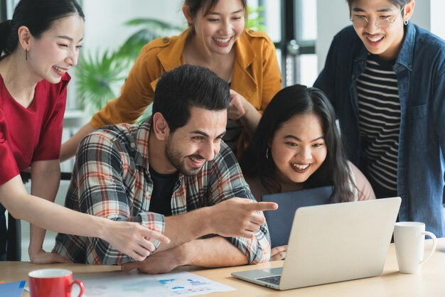 Trendiger asiatischer kreativer Hipster-Freund, der lächelt, während er bei einem zwanglosen Treffen mit einer Gruppe von Freunden sitzt, arbeiten an einem neuen Erfolgsprojekt, während sie digitale Laptops verwenden