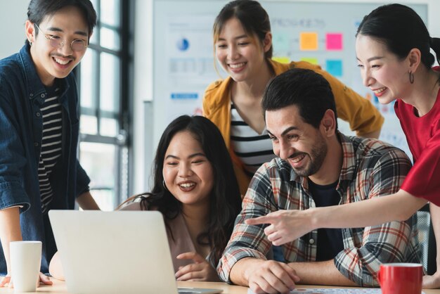 Trendiger asiatischer kreativer Hipster-Freund, der lächelt, während er bei einem zwanglosen Treffen mit einer Gruppe von Freunden sitzt, arbeiten an einem neuen Erfolgsprojekt, während sie digitale Laptops verwenden