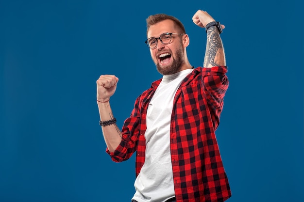 Trendiger Mann im karierten Hemd, der die Fäuste als Geste des Erfolgs hochhält und glücklich mit Gewinn auf blauem Hintergrund schreit. Hipster