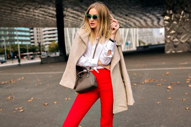 Trendiges Herbstmodeporträt der stilvollen jungen Frau, die nahe modernes Architekturgebäude aufwirft, das Hipster-Geschäftsoutfit und -mantel, Weinlese-Sonnenbrille, getönte Farben trägt.